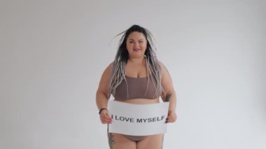 Çeşitli güzellik, çeşitlilik hoşgörü konsepti. İç çamaşırlı mutlu şişman bir kadın elinde pozitif bir vücut posteri tutuyor. Tatlandırıcıdaki şişman kız kameraya gülümsüyor..