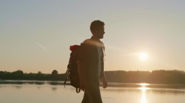 Gün doğumunda nehrin kenarında büyük bir sırt çantasıyla yürüyüş yapan genç bir adamın silueti. Yolculuğun tadını çıkaran bir adam. Kendine karşı zafer kazanıyorsun. Yürüyüş kavramı, sağlıklı yaşam tarzı, seyahat. Tek başına etkinlik