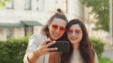 İki genç, gülümseyen hippi sarışın kadın akıllı telefondan kendi portresini çekiyor. Sokak geçmişinde çok şık kızlar var. Olumlu duygular ve aşk. LGBTQI, Gurur Etkinliği, LGBT Gurur Ayı