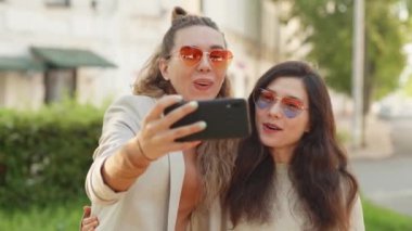 Olumlu duygular ve aşk. İki genç, gülümseyen hippi sarışın kadın akıllı telefondan kendi portresini çekiyor. Sokak geçmişinde çok şık kızlar var. LGBTQI, Gurur Etkinliği, LGBT Gurur Ayı