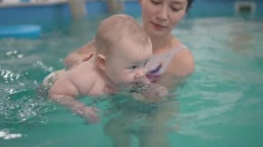 Siyah beyaz mayo giymiş mutlu anne ve bebek yüzme havuzunda eğlenerek suyun üstünde sörf yapıyor. Sağlıklı bir yaşam tarzı, aktif bir ebeveyn, çocuklarla kapalı havuzda yüzme dersleri.