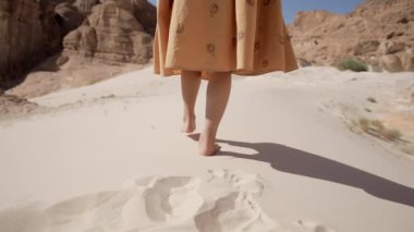 Yalnız kadın bacakları çölde yalın ayak kumda dolaşır ve geride yakın plan ayak izleri bırakır. Sarı elbiseli kız boş bir yerde tek başına yürüyor..
