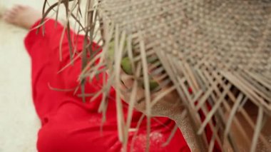 Alex kumsalda hindistan cevizini içki olarak tutan kadın elleri yakın plan. Uzun kırmızı elbiseli ve büyük hasır şapkalı bir kız beyaz kumda oturuyor ve meyvelerden hindistan cevizi sütü içiyor. Kavram