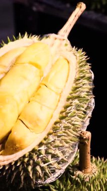Durian meyvesinin yarısı kesilmiş ve Asya 'da popüler tropikal lezzetlerle süslenmiş, dikenli kabuklu ve kremalı, egzotik meyve kültürü ve sokak piyasası titreşimleri gösteren bir meyve tezgahında sergileniyor. Yüksek kalite 4k görüntü