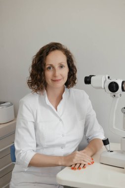 Kırk yaşlarında, beyaz önlüklü kıvırcık saçlı kadın doktor, klinikte profesyonel tıbbi bakım ve göz sağlığı değerlendirmesi yapan göz hekimliği cihazını kullanarak hastanın gözlerini inceliyor ve analiz ediyor. Yüksek kalite fotoğraf