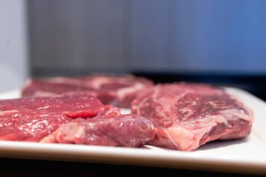 Büyük ve lezzetli bir ızgara biftek eti ve taze ev yapımı yemekleri tatmak için harika bir yoldur. Fotoğrafta beyaz bir tabakta üç büyük parça çiğ et görünüyor. Çerçeve aşağıdan çekilmiş. Yüksek kalite fotoğraf