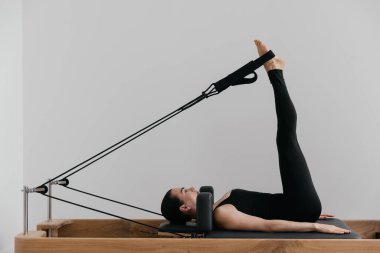 Modern bir stüdyoda siyah mayo giyen beyaz Avrupalı bir kadın olarak kullanılan pilates reformist eğitim makinesi, fitness yaşam tarzı dengesini gösteren beyaz bir duvara karşı kontrollü pilates egzersizleri yapıyor. Yüksek kalite fotoğraf