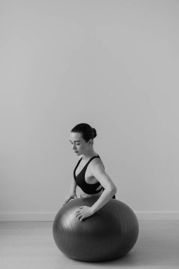 Stüdyoda kadın pilates topuyla antrenman yaparken çekilmiş siyah beyaz bir fotoğraf.