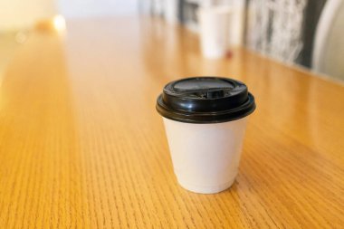 Siyah plastik kapaklı, tek kullanımlık beyaz kahve fincanı. Sarı ahşap masa üzerinde duruyor. Arka planı bulanık.