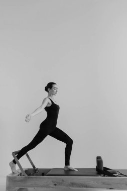 Zayıf bir kadının, odaklanmış fitness oturumunda kontrollü hareket, güç ve esneklik gösteren özel Pilates Reform Makinesi üzerinde egzersiz yaparken çekilmiş siyah ve beyaz fotoğrafı.