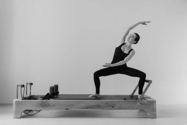 Zayıf bir kadının, odaklanmış fitness oturumunda kontrollü hareket, güç ve esneklik gösteren özel Pilates Reform Makinesi üzerinde egzersiz yaparken çekilmiş siyah beyaz bir fotoğrafı. Yüksek kalite fotoğraf