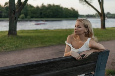 Güzel sarışın kadın yeşil şehir parkında nehir kenarındaki bankta oturuyor.