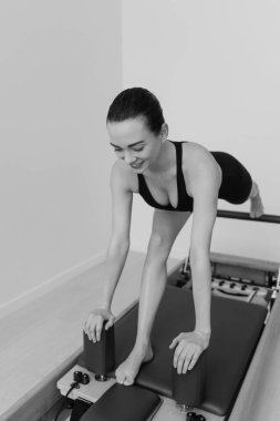 Zayıf bir kadının, odaklanmış fitness oturumunda kontrollü hareket, güç ve esneklik gösteren özel Pilates Reform Makinesi üzerinde egzersiz yaparken çekilmiş siyah beyaz bir fotoğrafı. Yüksek kalite fotoğraf