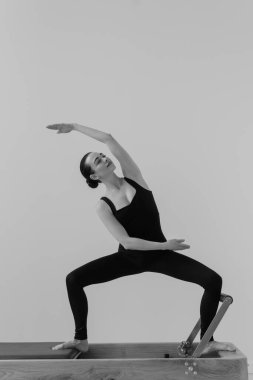 Zayıf bir kadının, odaklanmış fitness oturumunda kontrollü hareket, güç ve esneklik gösteren özel Pilates Reform Makinesi üzerinde egzersiz yaparken çekilmiş siyah ve beyaz fotoğrafı.