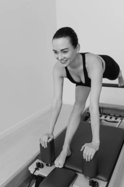 Zayıf bir kadının, odaklanmış fitness oturumunda kontrollü hareket, güç ve esneklik gösteren özel Pilates Reform Makinesi üzerinde egzersiz yaparken çekilmiş siyah beyaz bir fotoğrafı. Yüksek kalite fotoğraf