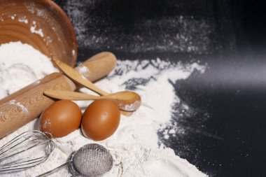 Ekranın solunda koyu bir yüzey üzerinde beyaz un yığını var. Yakınlarda yemek pişirmek için mutfak aletleri ve iki tavuk yumurtası var.. 