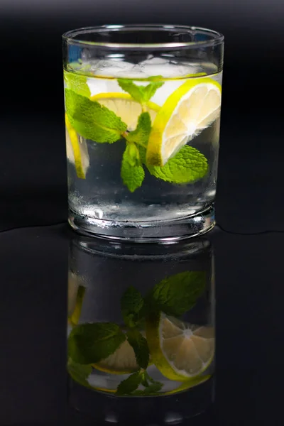 In einem Glas Wasser, Eiswürfeln, einem Zweig Minze und einer Zitronenscheibe. Spiegelung von der schwarzen Oberfläche des Tisches. — Stockfoto