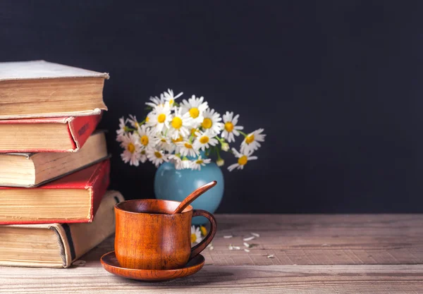 Una pila de libros antiguos de época sobre una mesa de madera, de pie junto a un jarrón azul con un ramo de margaritas y una taza de bebida caliente. País bodegón . —  Fotos de Stock
