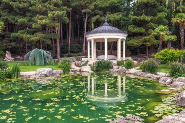 Bahçedeki yeşil göletin yanındaki beyaz köşkte yaz günü yansıması var. Kozalaklı ormandaki oyuk mimarisi. Kırım,