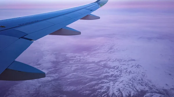 Air plane flies among the clouds over snowy capped mountains. Winter aerial landscape on purple sunset, view of the wing from the aircraft window.