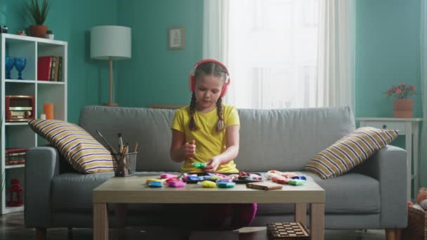 Menina pequena em fones de ouvido sem fio é rolamento de plasticina — Vídeo de Stock