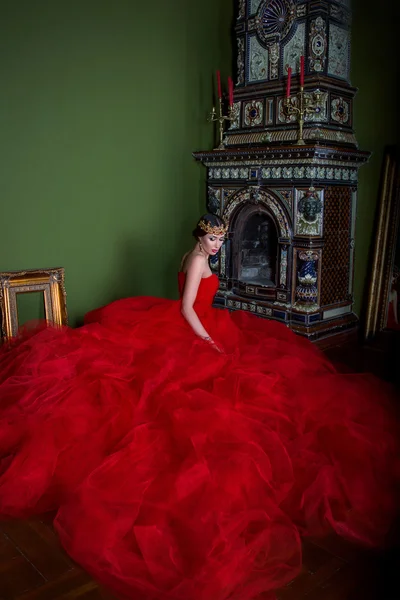 Beautiful girl in long red dress and in royal crown near fireplace — Stock Photo, Image