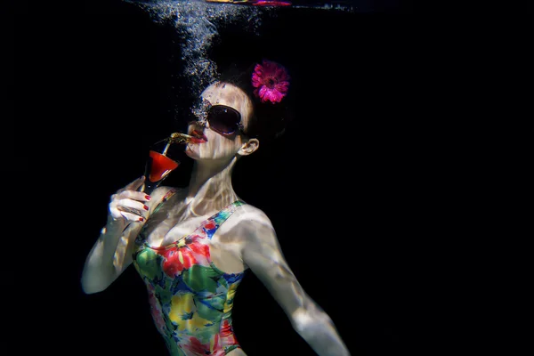 Όμορφο κορίτσι με ένα πολύχρωμο κομψό μαγιό και γυαλιά ηλίου και ροζ ζέρμπερες στο κεφάλι με ένα κοκτέιλ στο χέρι κάτω από το νερό στην πισίνα — Φωτογραφία Αρχείου