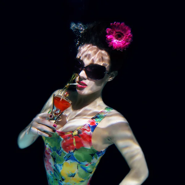 Красива дівчина в барвистому стильному купальнику і сонцезахисних окулярах і рожевій гербері в голові з коктейлем в руці під водою в басейні — стокове фото