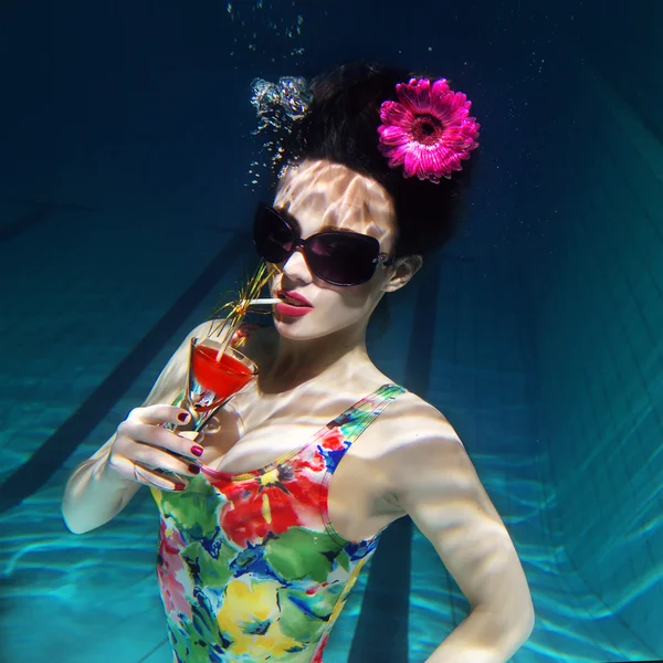 Mooi meisje in een kleurrijke, stijlvolle zwembroek en zonnebrillen en roze gerbera in het hoofd met een cocktail in haar hand onder water in het zwembad — Stockfoto
