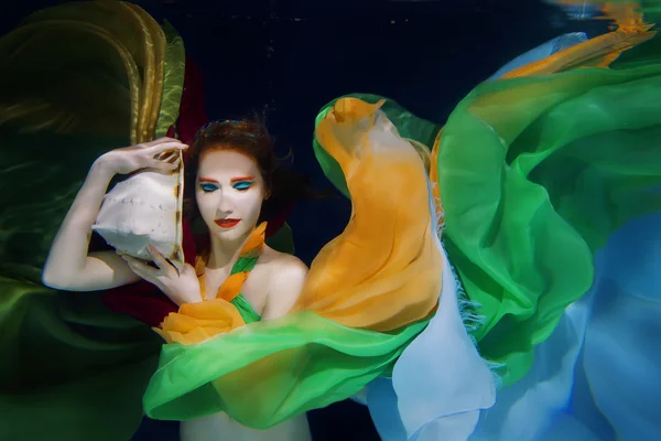 Chica de pelo rojo con concha de mar grande en ropa de colores en el fondo oscuro bajo el agua — Foto de Stock