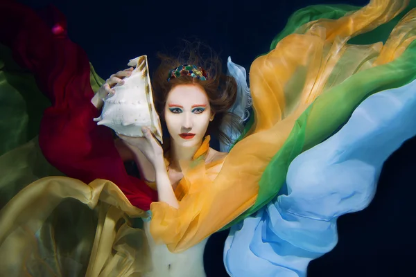 Chica de pelo rojo con concha de mar grande en ropa de colores en el fondo oscuro bajo el agua — Foto de Stock