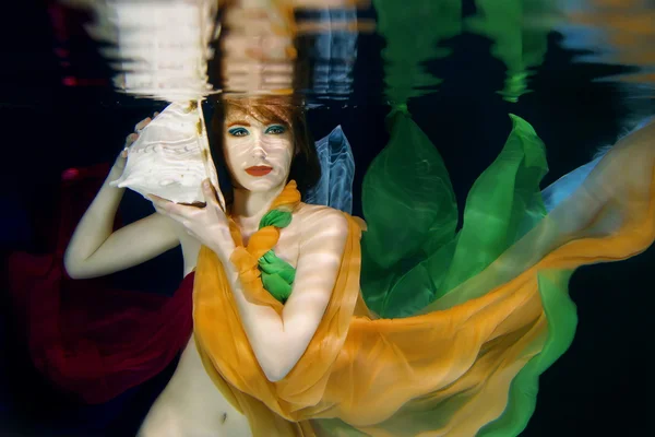 Chica de pelo rojo con concha de mar grande en ropa de colores en el fondo oscuro bajo el agua — Foto de Stock