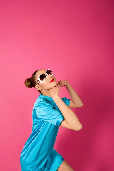 Porträt einer jungen schönen Frau in blauem Seidenkleid und weißer Sonnenbrille vor rosa Hintergrund — Stockfoto