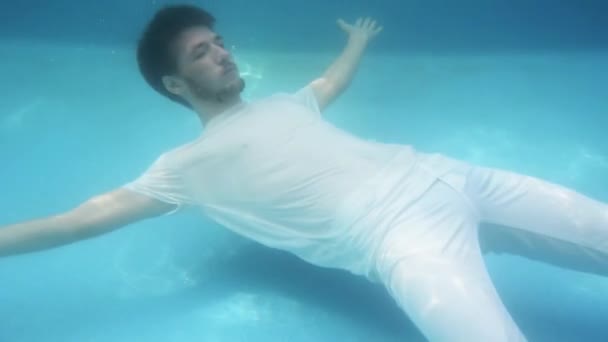 Junger Mann in weißem T-Shirt und weißer Hose taucht im Schwimmbad unter Wasser und schwimmt an die Oberfläche, taucht auf. — Stockvideo
