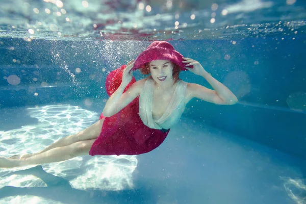 Lachende jonge mooie witte vrouw in jurk en roze hoed onder water in het zwembad — Stockfoto