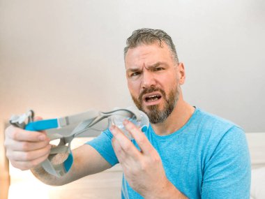 Mutsuz, şok olmuş, kronik solunum sorunu olan bir adam yatak odasında CPAP makinesini kullanarak şaşırdı. Sağlık, CPAP, uyku apnesi terapisi, horlama konsepti
