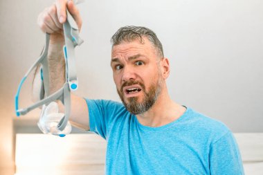 Mutsuz, şok olmuş, kronik solunum sorunu olan bir adam yatak odasında CPAP makinesini kullanarak şaşırdı. Sağlık, CPAP, uyku apnesi terapisi, horlama konsepti