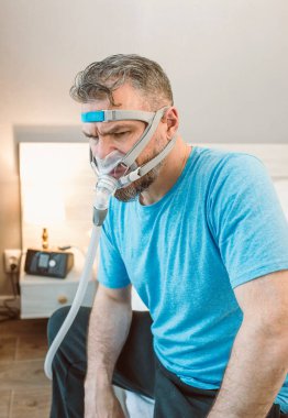 Mutsuz, şok olmuş, kronik solunum sorunu olan bir adam yatak odasında CPAP makinesini kullanarak şaşırdı. Sağlık, CPAP, uyku apnesi terapisi, horlama konsepti