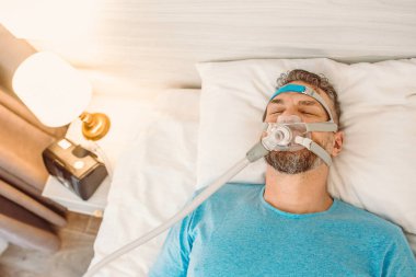 Kronik solunum sorunu olan uyuyan adam yatakta CPAP makinası kullanmayı düşünüyor. Sağlık, obstrüktif uyku apnesi terapisi, CPAP, horlama konsepti