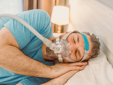 Kronik solunum sorunu olan uyuyan adam yatakta CPAP makinası kullanmayı düşünüyor. Sağlık, obstrüktif uyku apnesi terapisi, CPAP, horlama konsepti