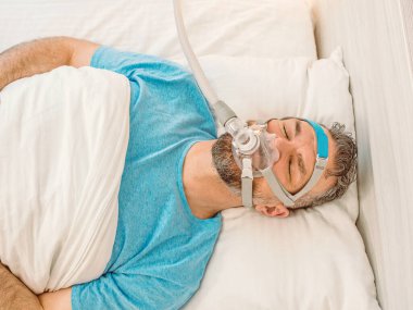 Kronik solunum sorunu olan uyuyan adam yatakta CPAP makinası kullanmayı düşünüyor. Sağlık, obstrüktif uyku apnesi terapisi, CPAP, horlama konsepti