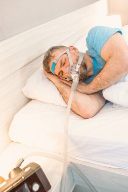Kronik solunum sorunu olan uyuyan adam yatakta CPAP makinası kullanmayı düşünüyor. Sağlık, obstrüktif uyku apnesi terapisi, CPAP, horlama konsepti