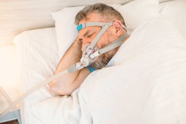 Kronik solunum sorunu olan uyuyan adam yatakta CPAP makinası kullanmayı düşünüyor. Sağlık, obstrüktif uyku apnesi terapisi, CPAP, horlama konsepti