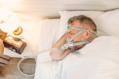 Kronik solunum sorunu olan uyuyan adam yatakta CPAP makinası kullanmayı düşünüyor. Sağlık, obstrüktif uyku apnesi terapisi, CPAP, horlama konsepti