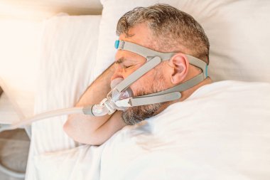 Kronik solunum sorunu olan uyuyan adam yatakta CPAP makinası kullanmayı düşünüyor. Sağlık, obstrüktif uyku apnesi terapisi, CPAP, horlama konsepti