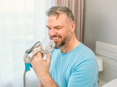 Kronik solunum sorunu olan olgun, mutlu bir adam yatak odasındaki yatakta oturan CPAP makinesini kullanmayı düşünüyor. Sağlık, CPAP, horlama konsepti