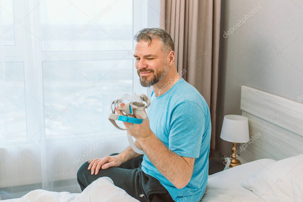 Mature happy man with chronic breathing issues considers sitting on the bed in bedroom with CPAP machine. Healthcare, CPAP, snoring concept