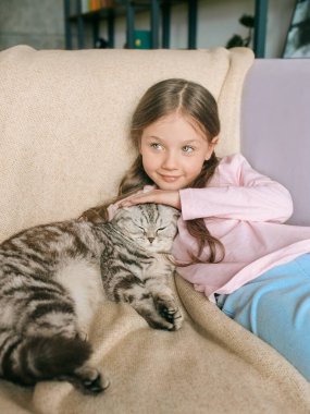 Sevimli, sevimli, gülümseyen kız evdeki kanepede İskoç gri kedisine sarılıyor. Evcil hayvanlar, arkadaşlık, ev, mutluluk konsepti 