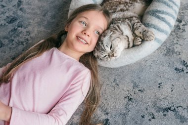 Sevimli, sevimli, gülümseyen kız evde halıda İskoç gri kedisiyle yatıyor. Evcil hayvanlar, arkadaşlık, ev, mutluluk konsepti 