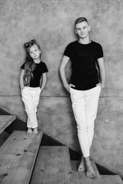 Siyah tişörtler ve beyaz pantolonlar içinde gri duvarın yanında duran beyaz pantolonlu beyaz erkek ve kız kardeşler. Aile, moda, akrabalar, ilişki ve çocuk kavramı.